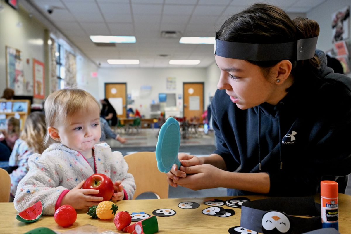 An adult is holding a mirror so that a toddler child, who is holding an apple, can see their face in it.